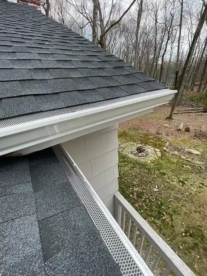 White seamless gutter with metal guard on asphalt shingle roof edge in wooded yard