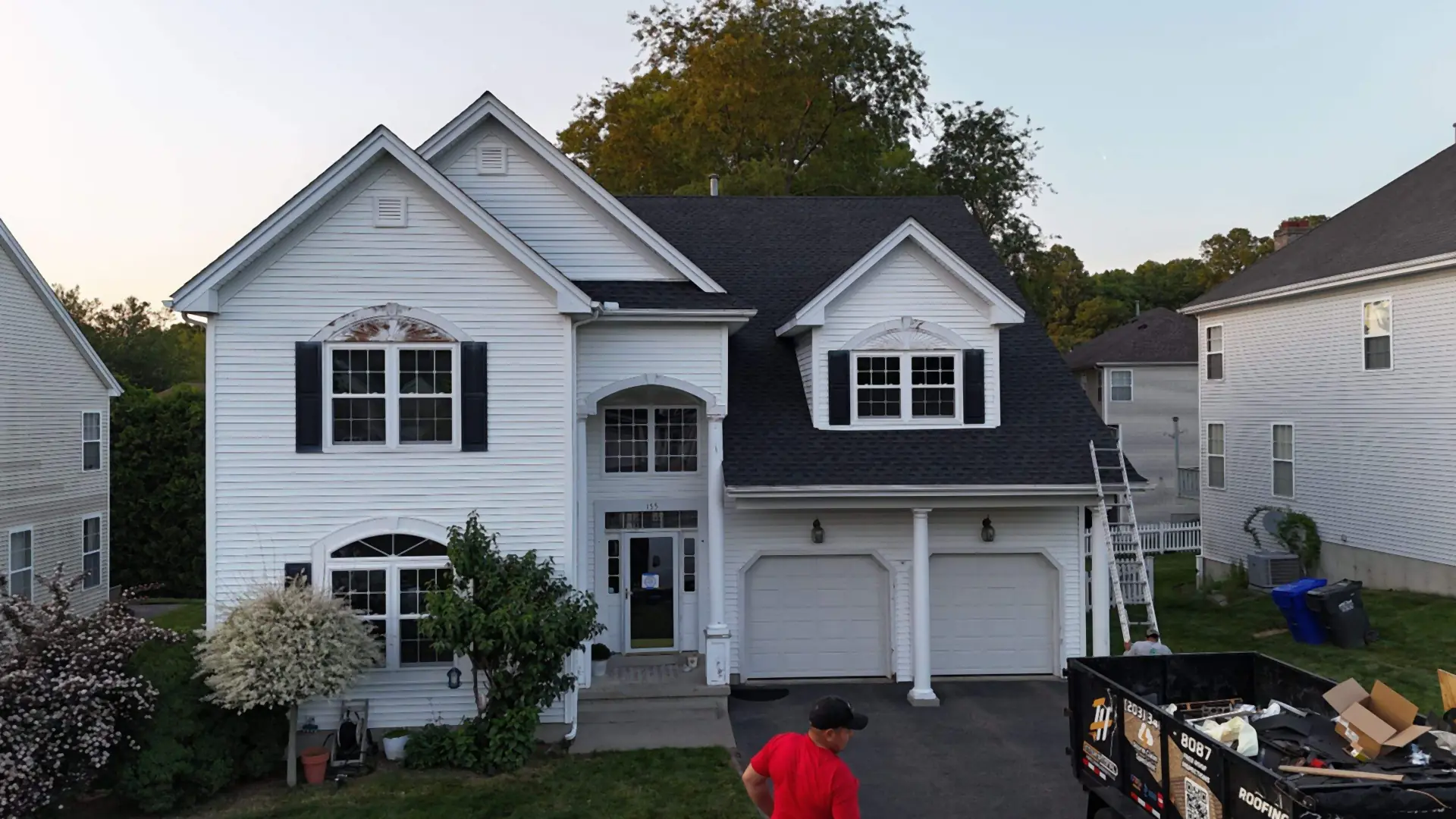 White two-story house with new dark roof, dumpster and worker during replacement